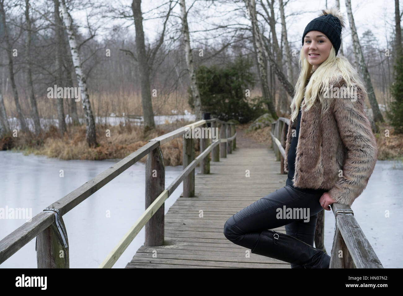 Schöne schwedische blonde Frau im schwarzen Kappe und schönes Outfit posiert auf Holzbrücke. Lächelnd, entspannt und glücklich. Stockfoto