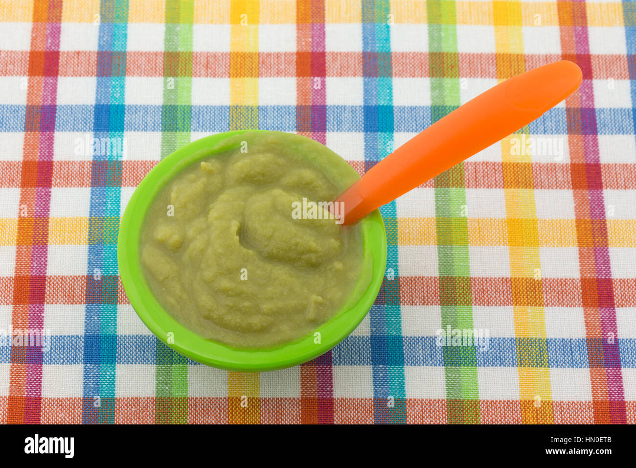 Eine Portion grüne Erbsen Babynahrung in einer kleinen Schüssel mit einem Plastiklöffel orange auf einem bunten Tischset. Stockfoto