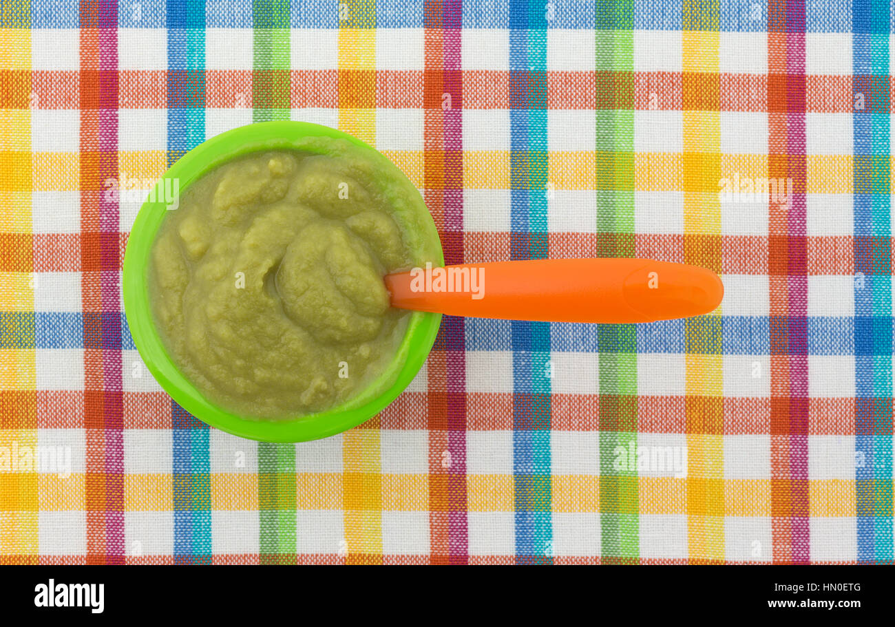 Draufsicht auf eine Portion grüne Erbsen Babynahrung in einer kleinen Schüssel mit einem Plastiklöffel orange auf einem bunten Tischset. Stockfoto