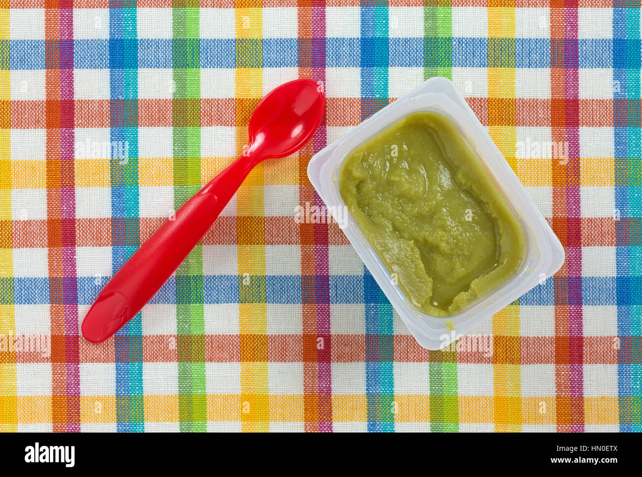 Draufsicht auf eine Portion grüne Erbsen Babynahrung in einem kleinen Plastikbehälter mit einem roten Löffel auf die Seite auf einem bunten Tischset. Stockfoto