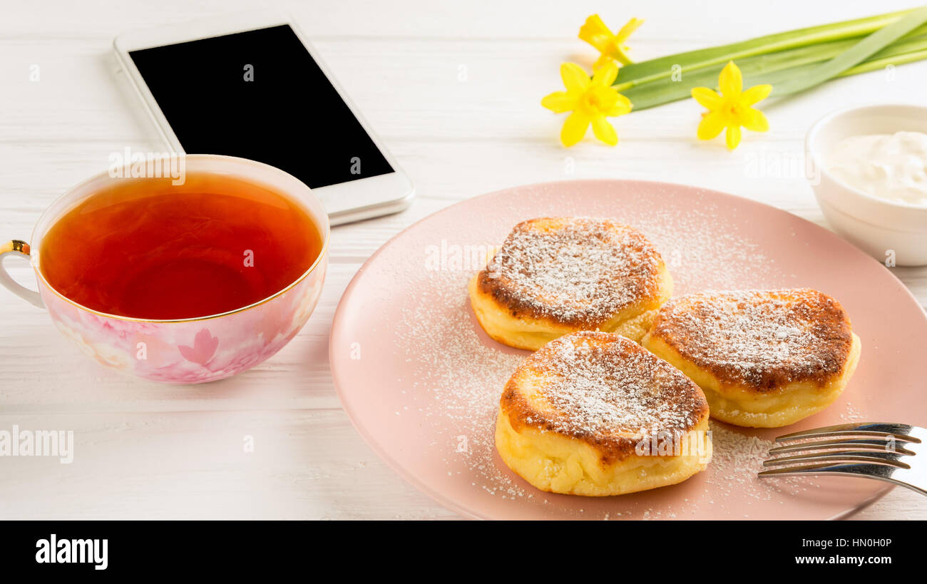 Käse-Pfannkuchen mit Puderzucker, schwarzer Tee, Blumen und Smartphone auf einem weißen Holztisch. Stockfoto