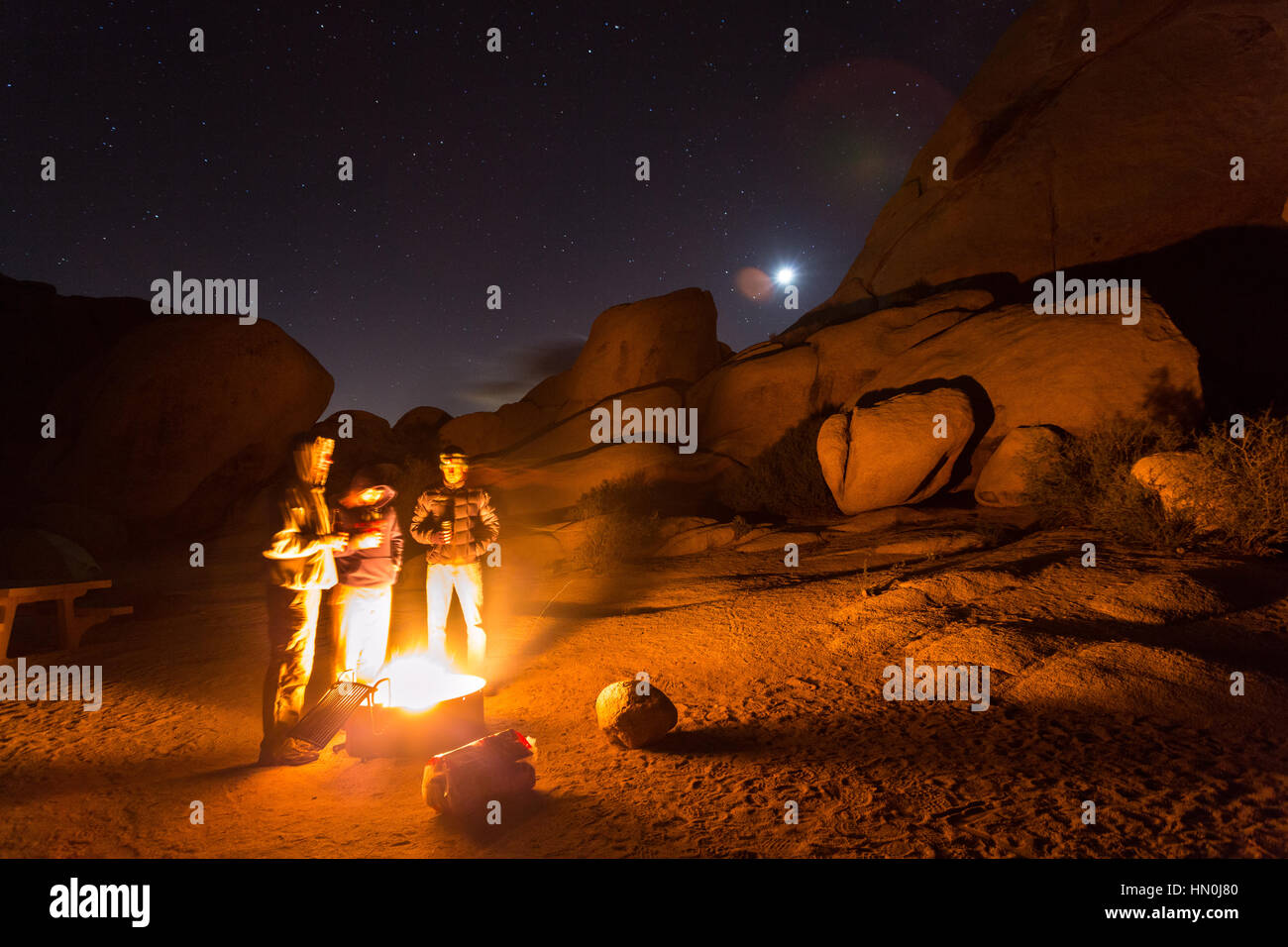 Drei Freunde stehen für Wärme an einem kalten und windigen Nacht im Joshua Tree National Park in der Nähe des Lagerfeuers. Stockfoto