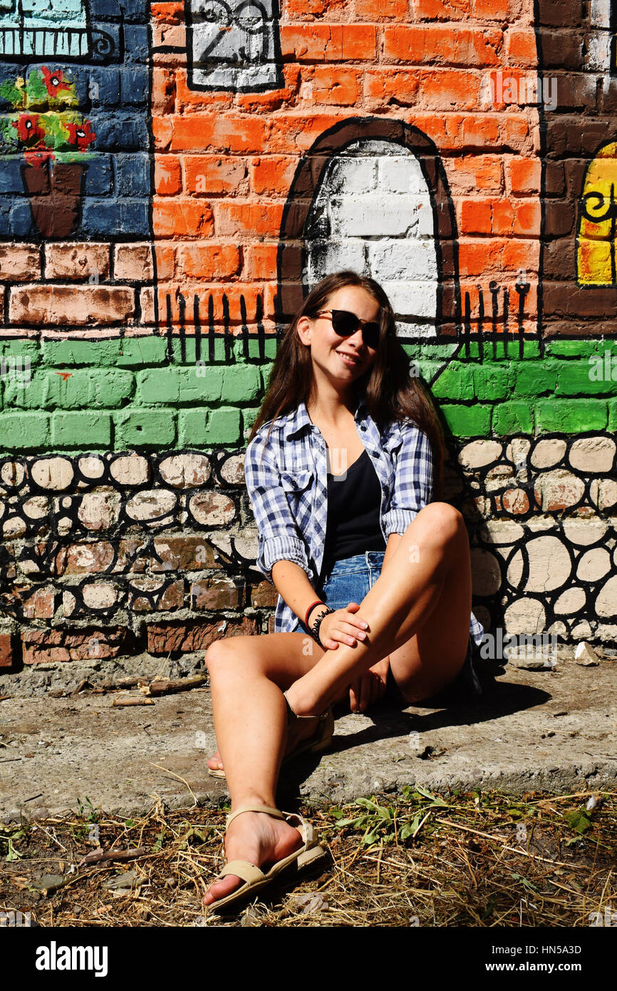 Porträt von schöne Teenager-Mädchen in Sonnenbrille weared auf karierten Hemd und Jeans Shorts, gegen eine Wand mit irgendein Element des Graffiti. Stockfoto