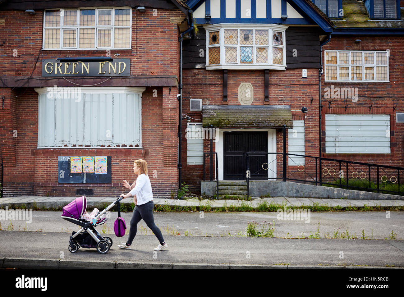 Junge Mutter schieben Kinderwagen letzten außen geschlossen und mit Brettern vernagelt Windows Grün Ende eine großen Wahrzeichen Kneipe in Burgage, South Manchester, England, UK, Stockfoto