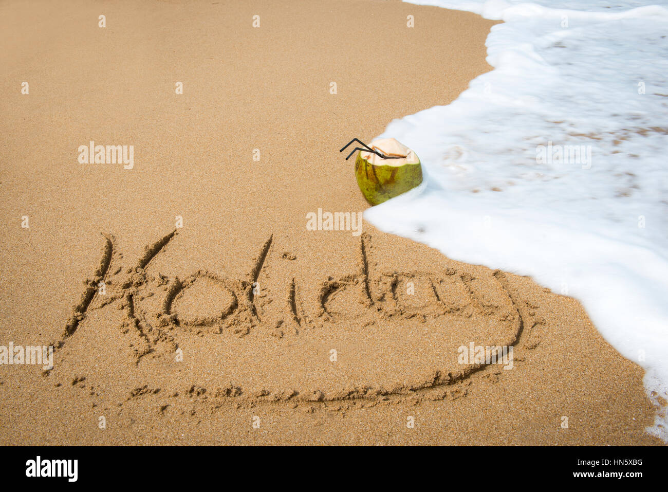 Das Wort Urlaub in den Sand mit einer Ozeanwelle darüber geschrieben war es und eine Kokosnuss Drink entfernt. Stockfoto