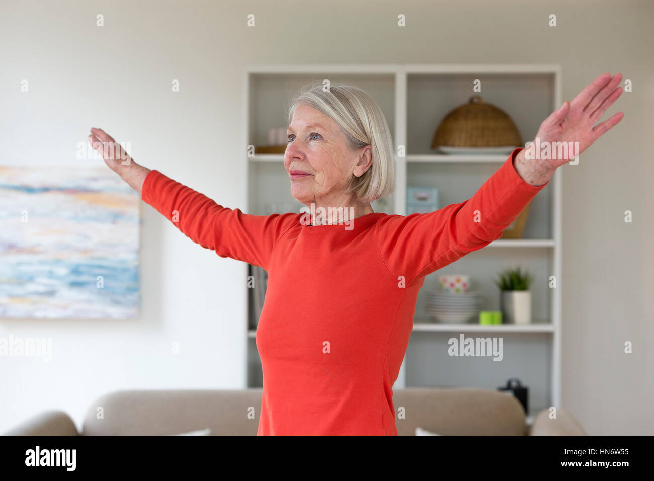 Ältere Frau Ausübung in ihrem Wohnzimmer. Sie hat ihre Arme ausgestreckt. Stockfoto