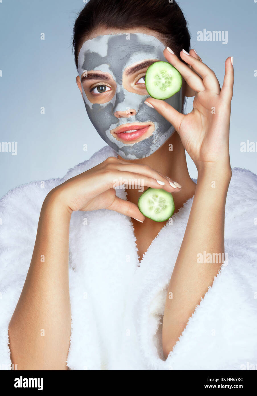 Schöne Mädchen in weißen Bademantel und mit Maske aus Ton. Foto von Brünette Frau mit Gurkenscheiben in der Nähe von ihrem Gesicht auf blauem Hintergrund. Pflege ihn Stockfoto