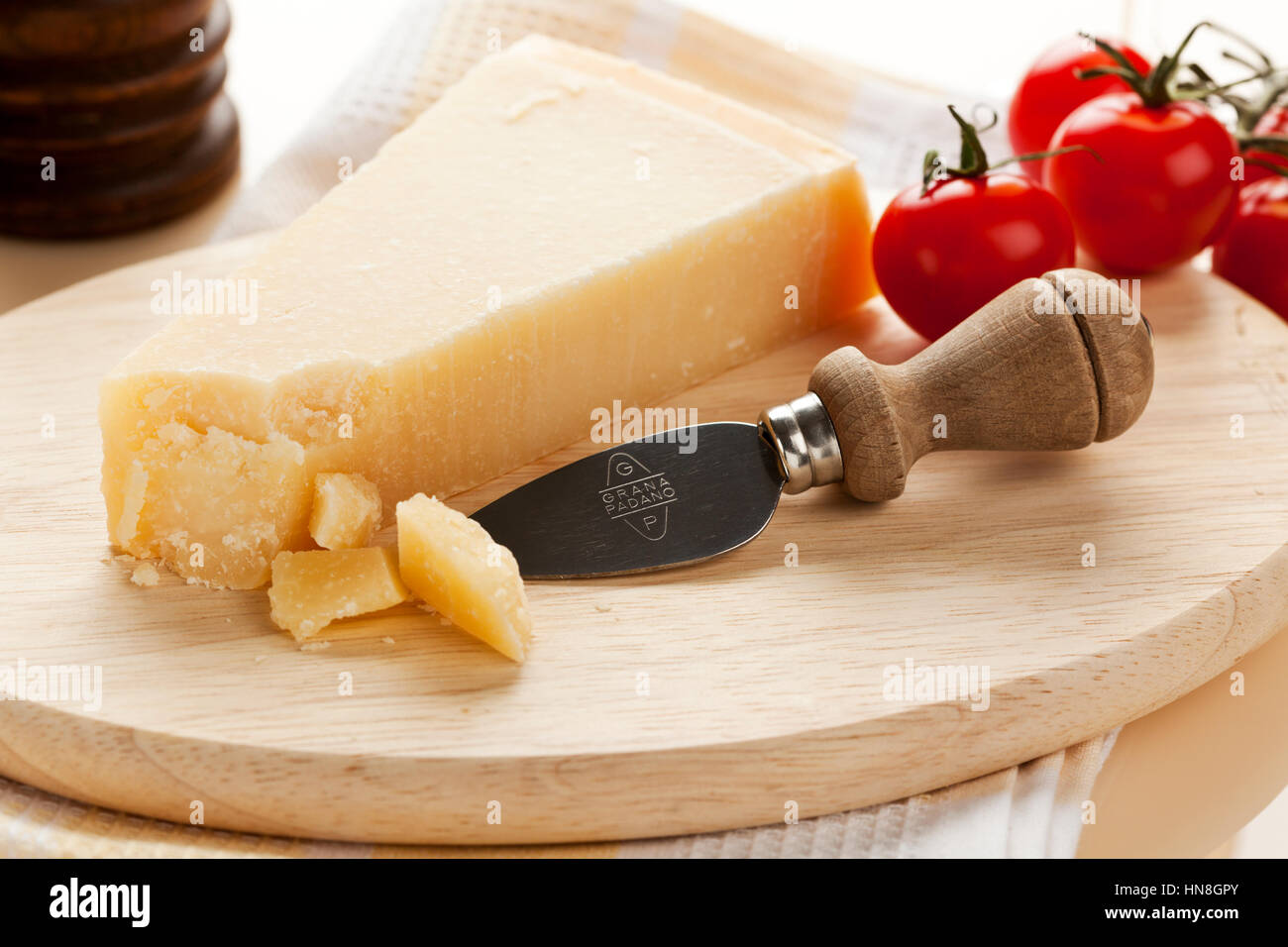 Grana Padano Käse und Messer mit Logo auf Holzplatte, Tomaten und Pfeffer Mühle im Hintergrund. Grana Padano ist ein harter chee Stockfoto