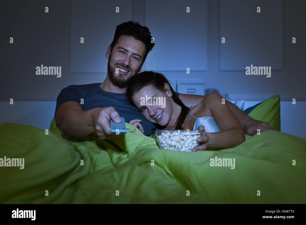 Glückliches Paar Filme im Fernsehen im Bett, der Mann hält eine Fernbedienung und die Frau ist Popcorn Essen Stockfoto