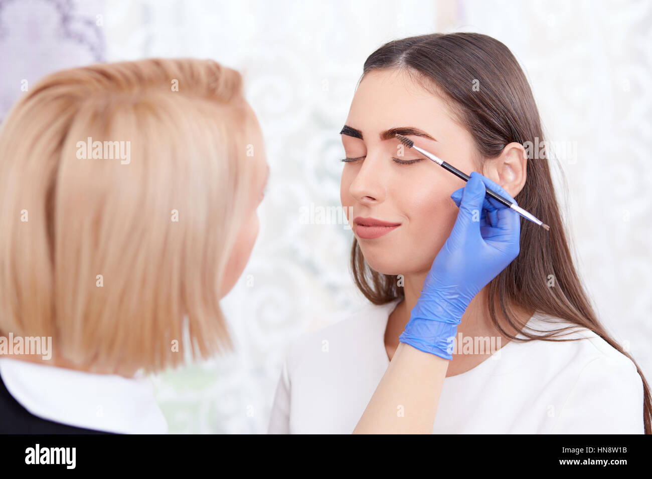 Porträt von professionellen blonde Kosmetikerin in blaue Handschuhe machen permanente Augenbrauen für Frau, mit Pinsel. Weiblichen Kunden mit geschlossenen Augen Lächeln Stockfoto