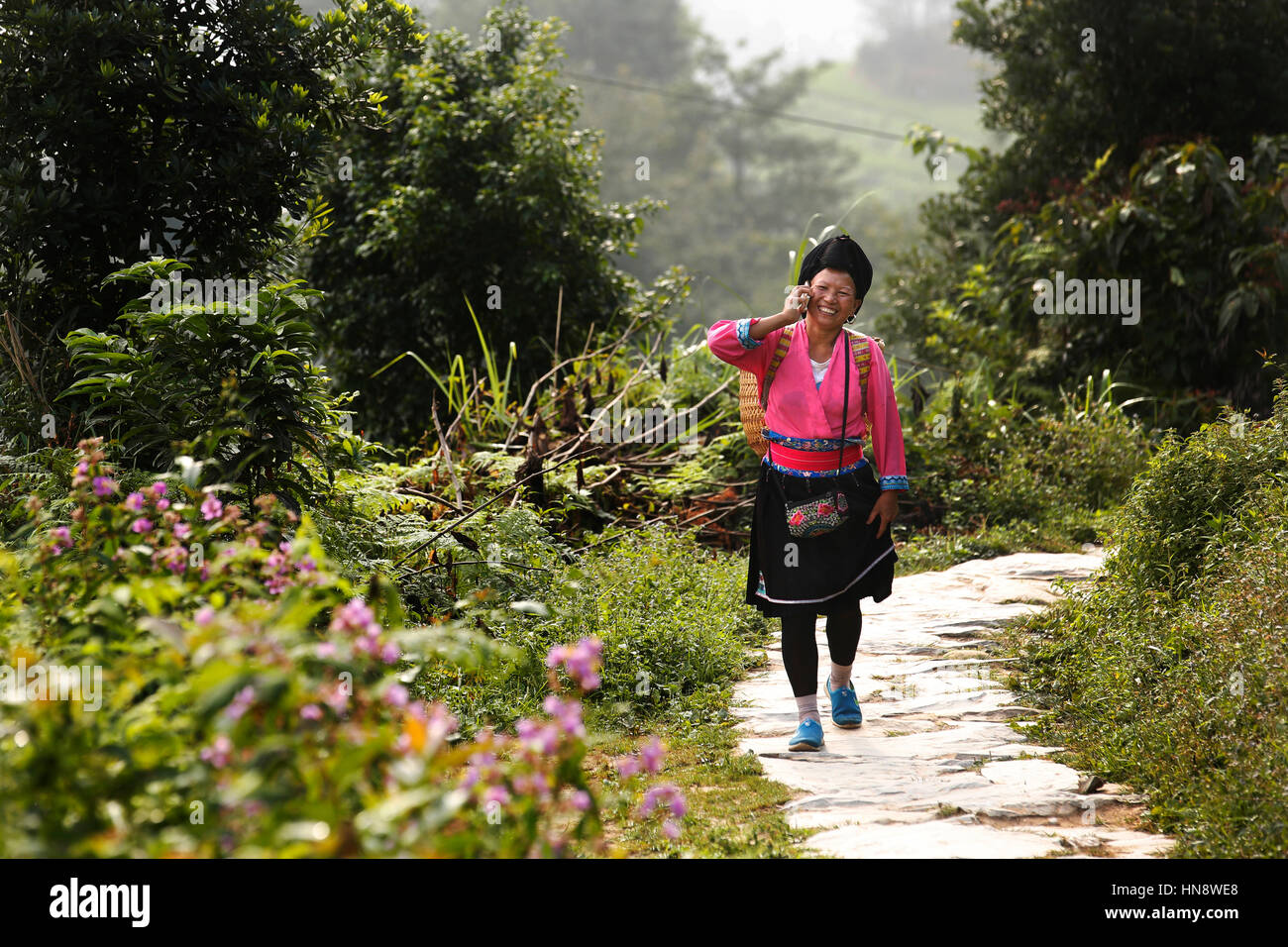 Eine Frau im traditionellen Yao Outfit spricht auf dem Handy wie geht sie entlang eines Pfades durch die Longji Rice-Terrassen in der Nähe von Guilin, Provinz Guangxi, Stockfoto
