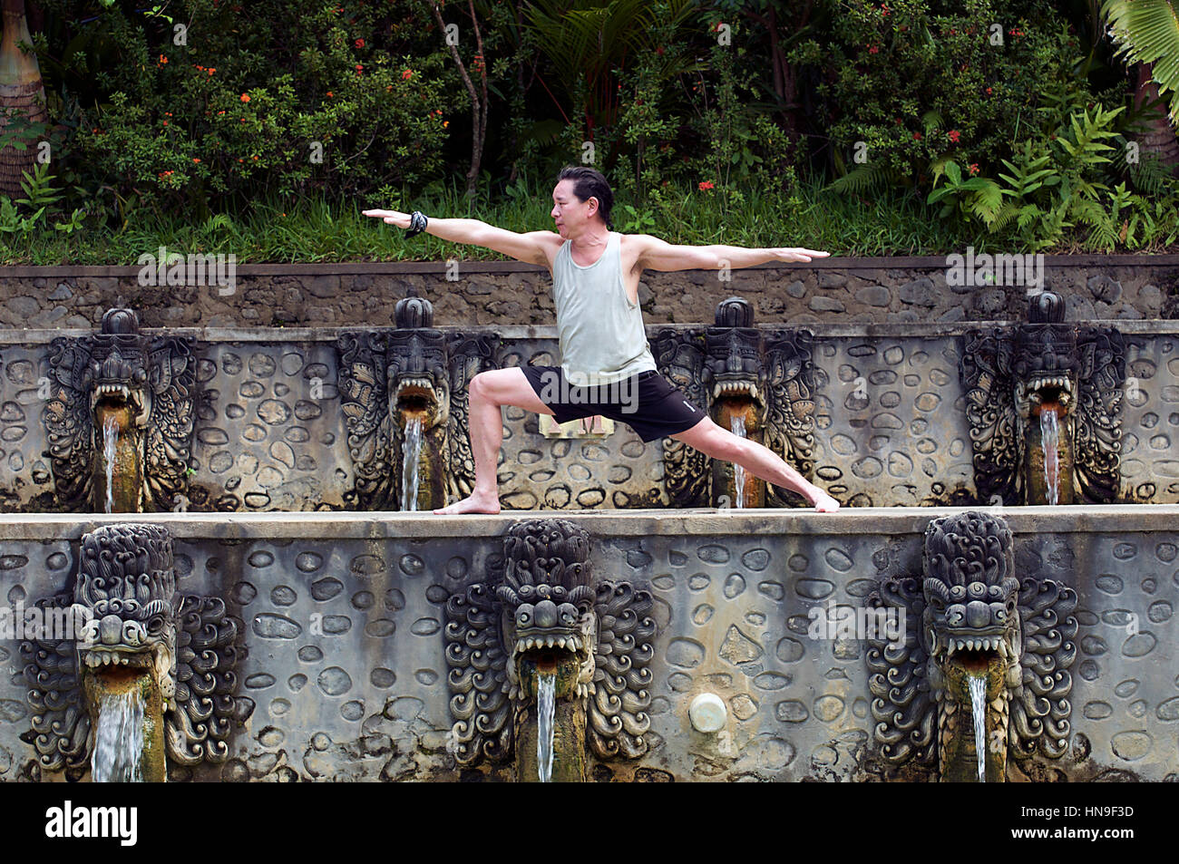 Asiatischen Mann mittleren Alters hält eine Krieger Hatha Yoga-Pose in Banjar Hot Springs Pools in Bali, Indonesien als Wasser von Drache-wie Geschöpfe Mund gießt Stockfoto