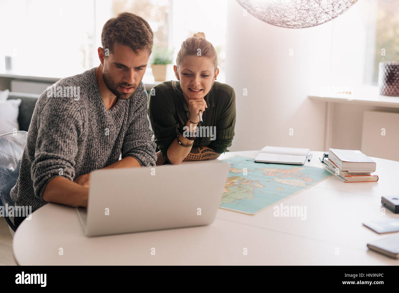 Junger Mann und Frau mit Weltkarte und Computer am Schreibtisch sitzen. Paar mit Technologie, um die Welt zu erkunden. Stockfoto
