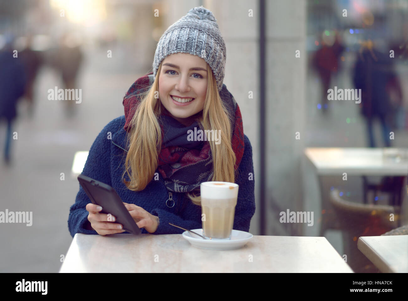 Junge Frau sitzen, einen Drink im Freien im Winter in ein Straßencafé sitzen halten ihre Handys oder kleine tablet schaut in die Kamera mit einem Stockfoto