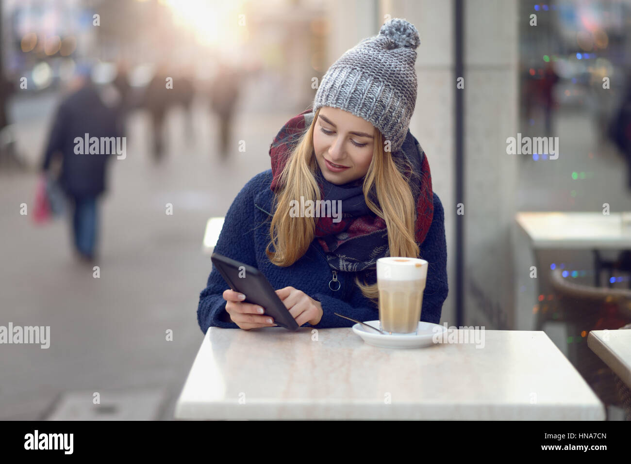 Junge Frau sitzen, einen Drink im Freien im Winter in ein Straßencafé sitzen halten ihre Handys oder kleine tablet schaut in die Kamera mit einem Stockfoto