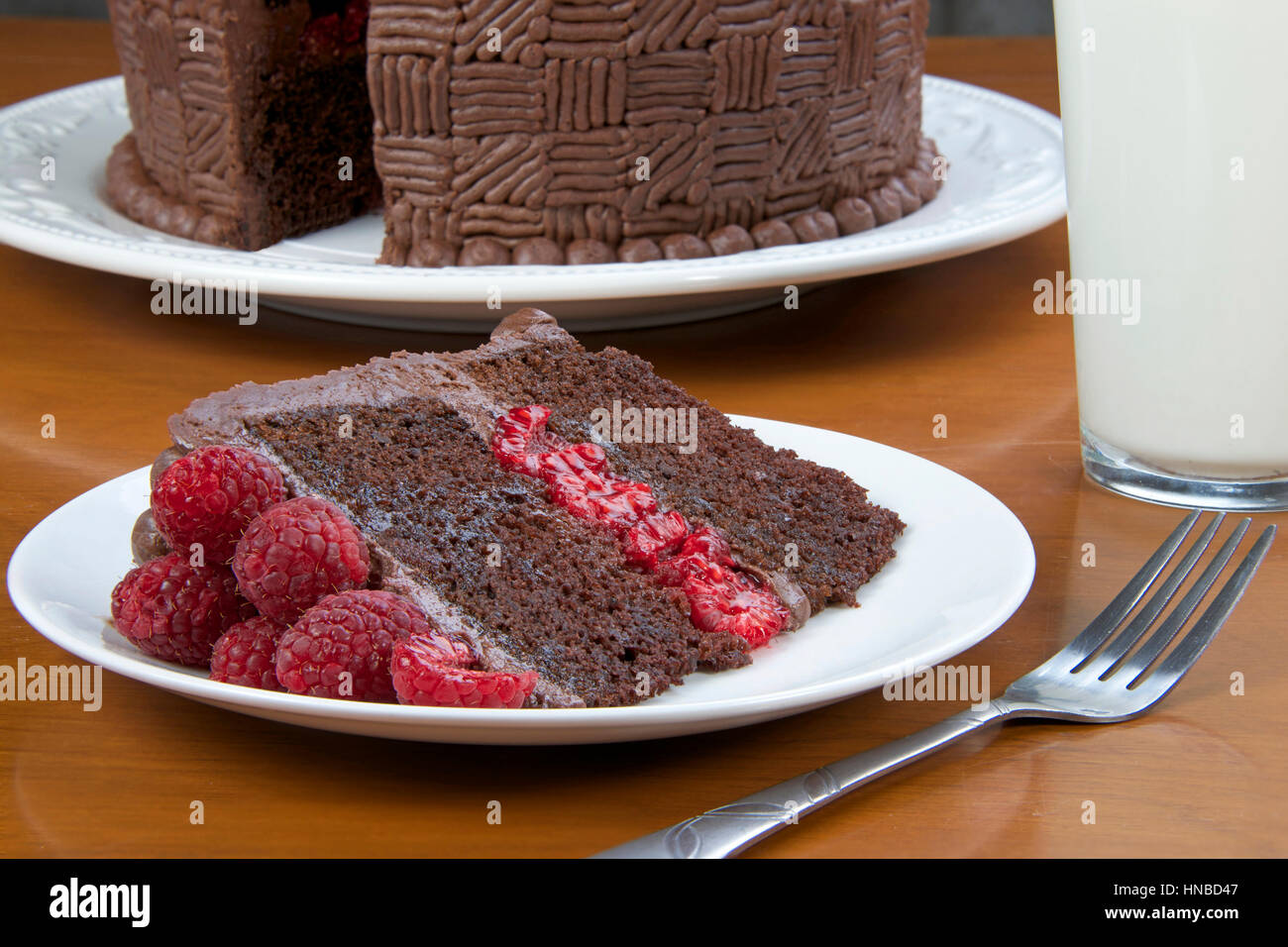 Stück Schokolade Himbeer Kuchen auf einem Teller mit einer Gabel, Glas Milch und ein Stück Kuchen schneiden im Hintergrund auf einem Holztisch. Stockfoto