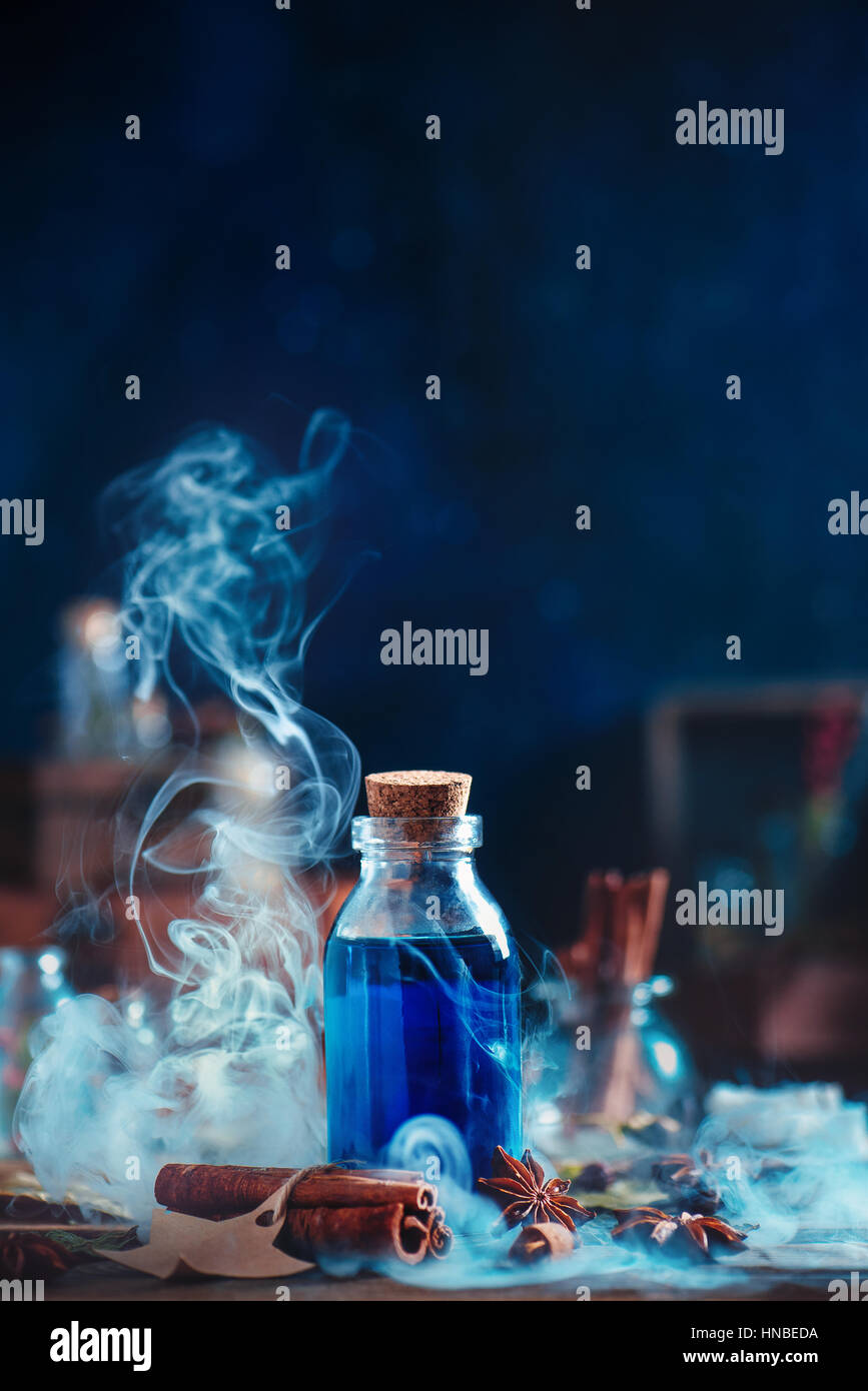 Stillleben mit Glasflasche blauer trank, Zimt, Kräuter und Dicke Wolken von steigenden Rauch auf dunkelblauem Hintergrund Stockfoto