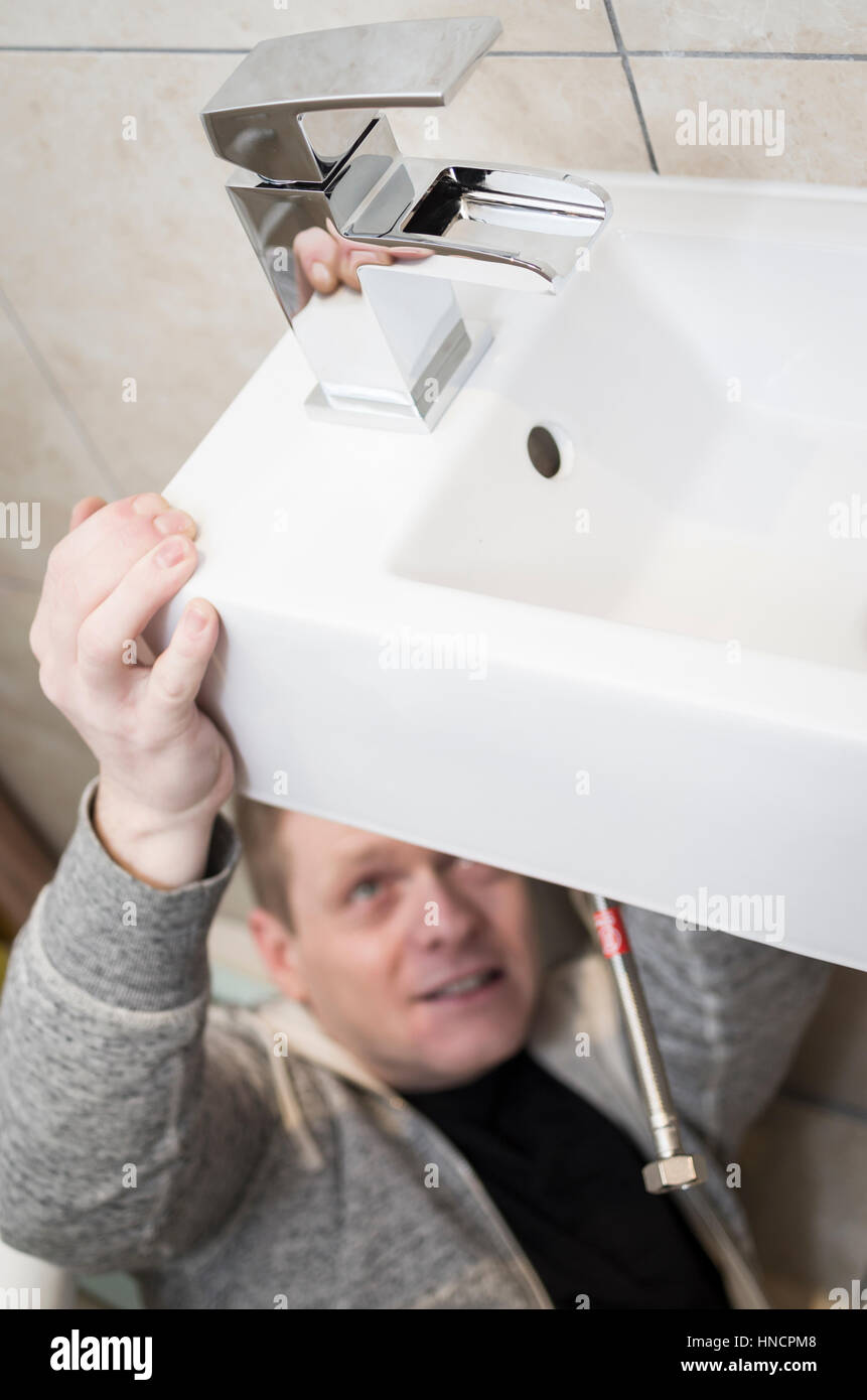 Junger Mann versucht tun es Yourself (DIY) Sanitär zu Hause unter einer Spüle Montage einen neuen Hahn mit lustigen Ausdruck auf seinem Gesicht Stockfoto