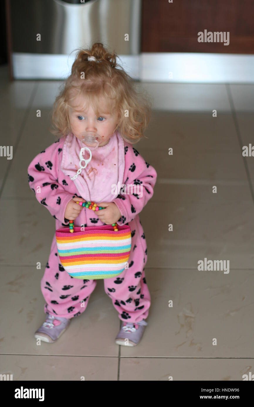 Süße lockigen blonden Kleinkind Mädchen Kind shopping Spielen mit einer Tasche und dodie im Mund trägt einen Strampler, spielen Konzept, Binky, Stockfoto