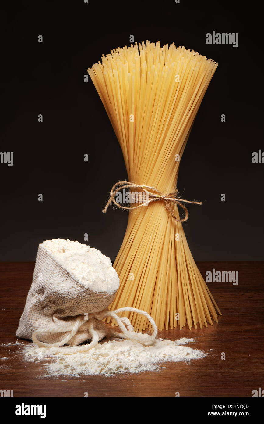 Trockene Spaghetti Nudeln gefesselt mit Seilen und Jute Beutel Mehl auf dunklen Holztisch auf grauem Hintergrund Stockfoto