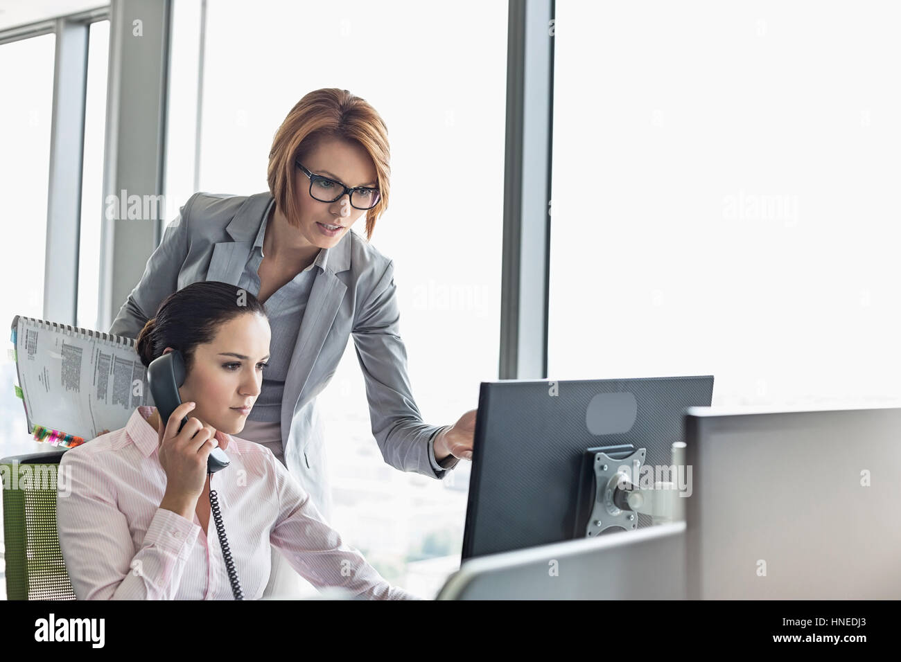 Junge Geschäftsfrau mit Festnetz-Telefon während Kollege zeigte auf Computer-Monitor im Büro Stockfoto
