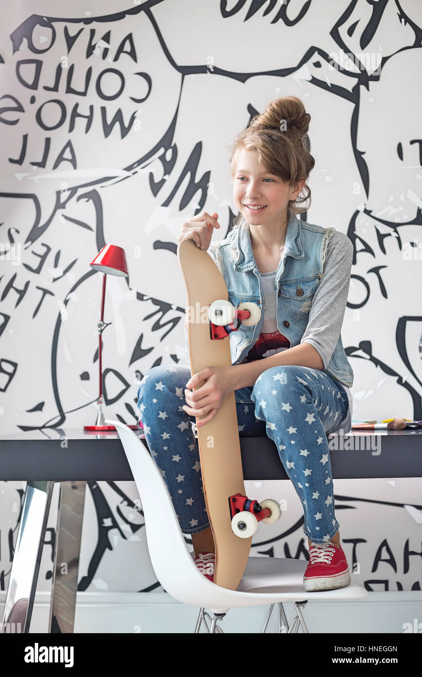 Mädchen mit Skateboard sitzt am Schreibtisch zu Hause in voller Länge Stockfoto