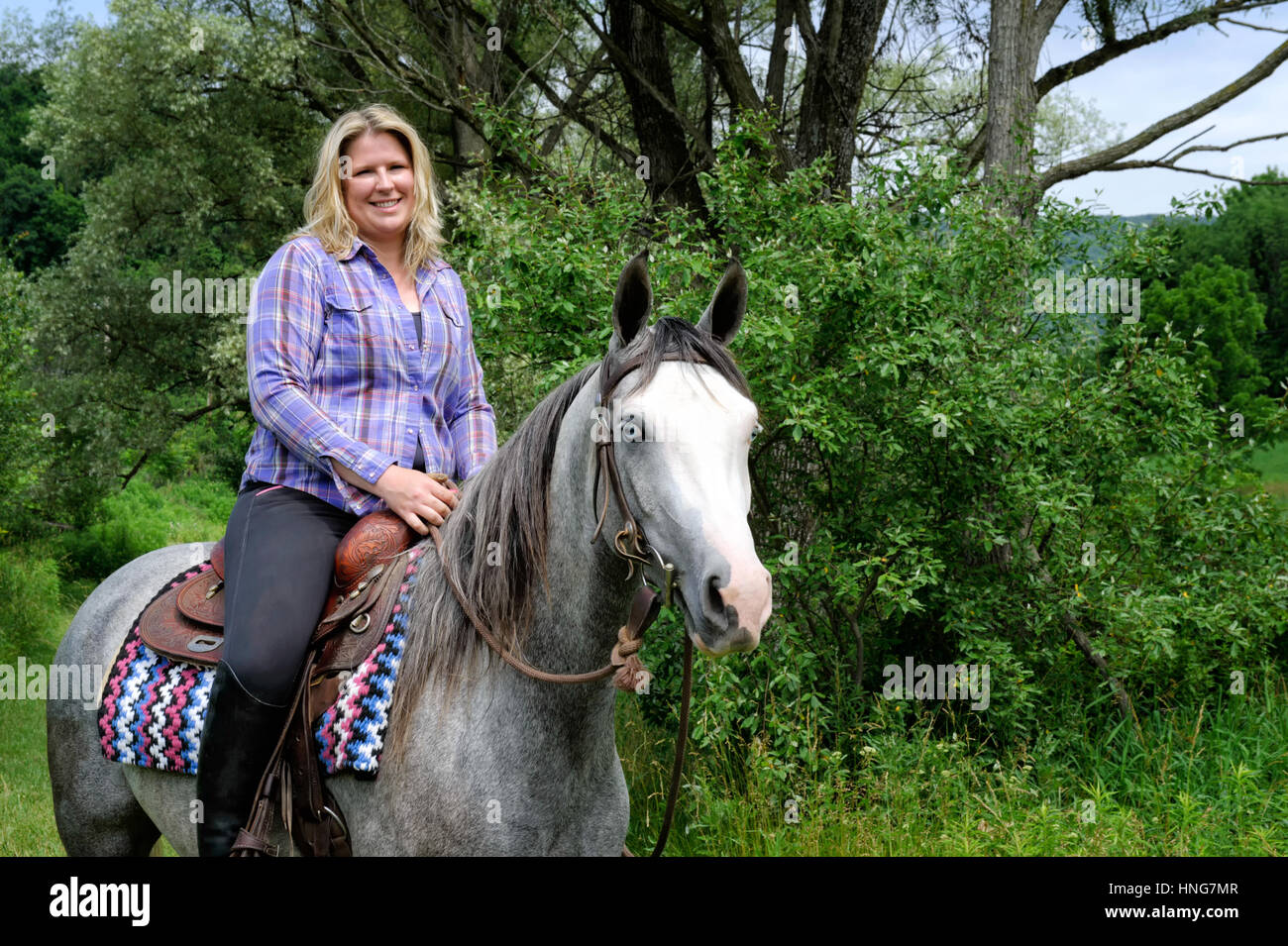 Blonde Frau in Alltagskleidung, montiert auf einem blauäugigen, White-faced, gescheckten Pferd Reiten western in Grasgrün Bereichen Sommer Laub. PA Stockfoto