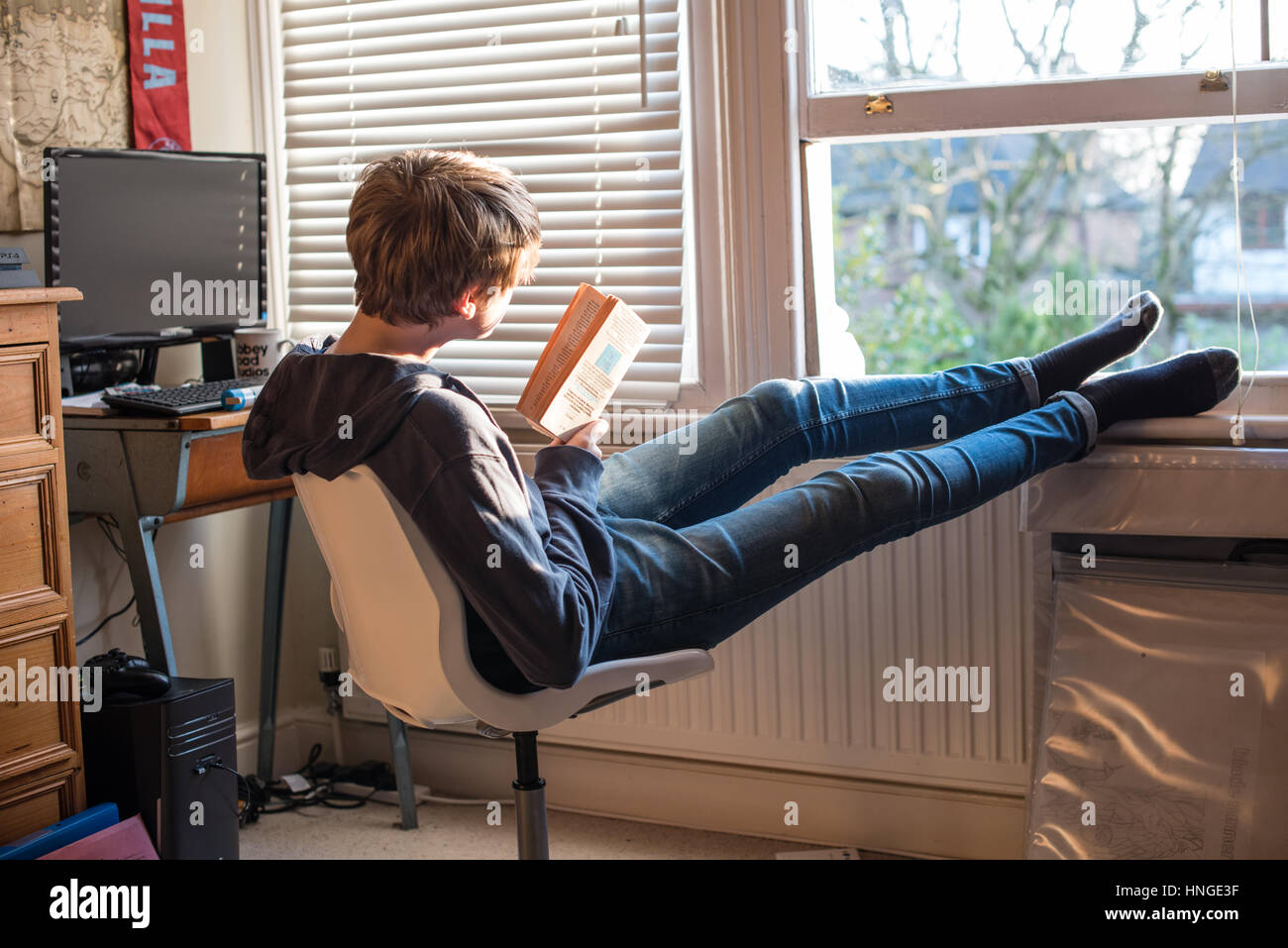 Weiße Teenager in Jeans und ein Schweiß-Top durch seine offene Schlafzimmerfenster mit seinen Füßen sitzend von seinem Schreibtisch lesen und Revision für GCSE Prüfungen Stockfoto