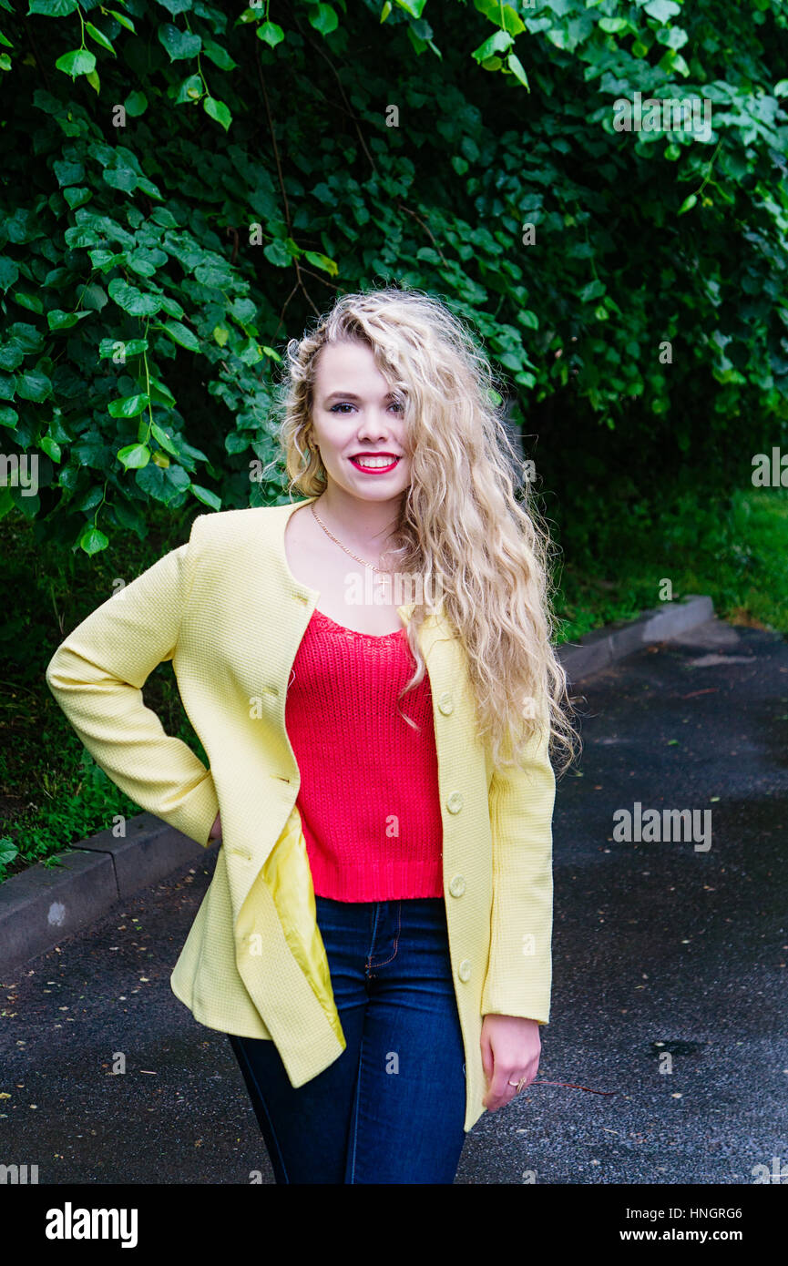 Charmante blonde Studentin posiert für die Kamera in einem grünen Park trägt eine gelbe Jacke, rote Jacke und jeans Stockfoto