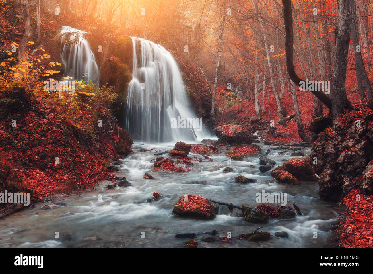 Wasserfall. Bunte Landschaft mit wunderschönen Wasserfall am Bergfluss im Wald mit rotem Laub bei Sonnenuntergang im Herbst. Bäume mit roten Blättern. St. Stockfoto