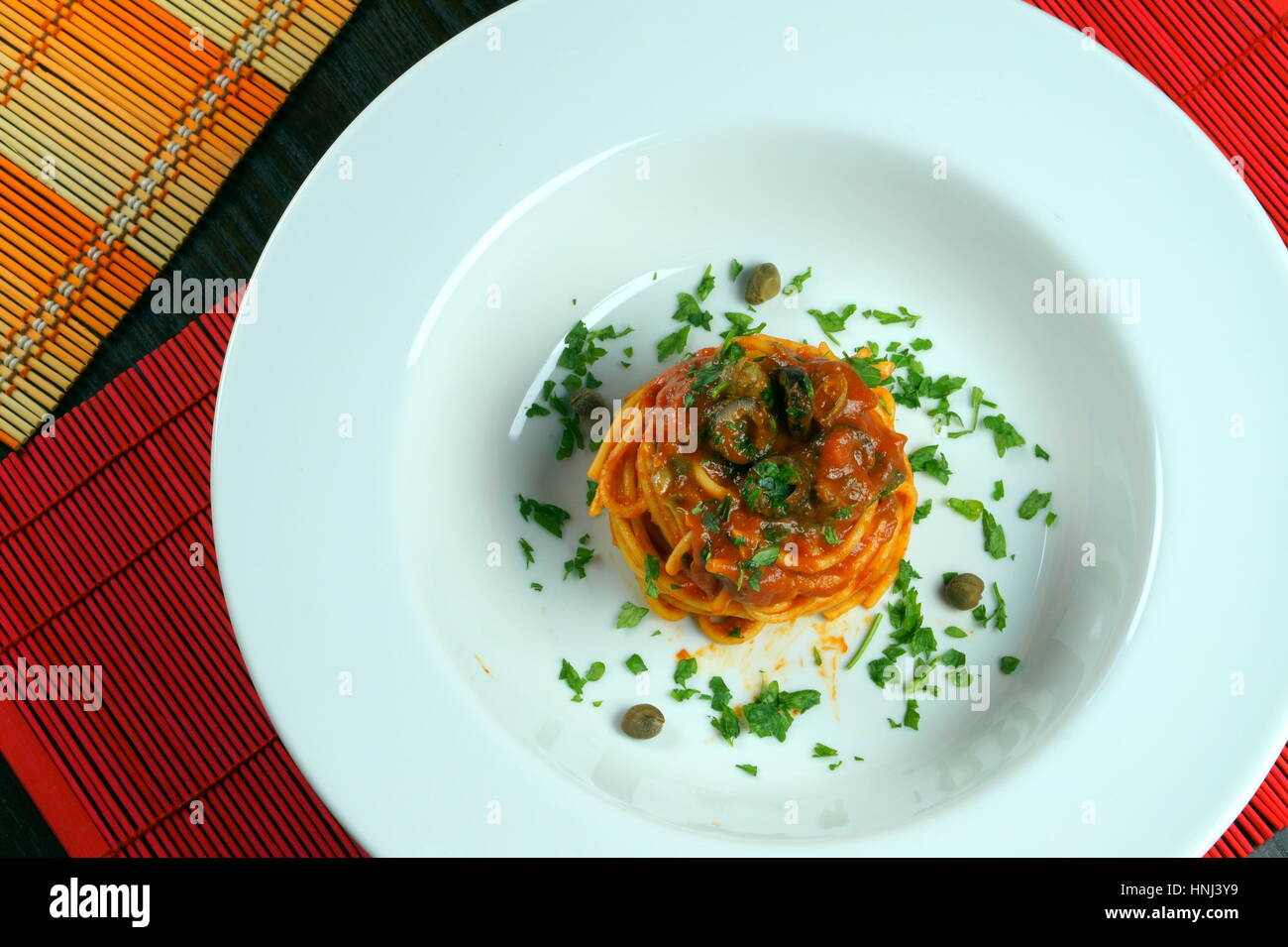 Spaghetti Puttanesca auf bunte Tischsets liegen auf Holztisch - italienisches Rezept mit Knoblauch, Olivenöl, Sardellen, Oliven, Tomaten, Petersilie, Kapern Stockfoto