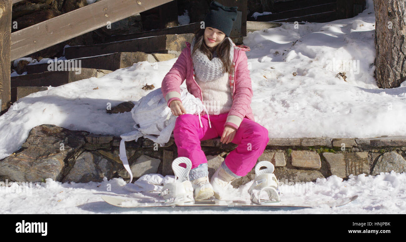 Hübsche junge Asiatin in rosa Schneeanzug entspannend auf Skiurlaub saß auf Mauer. Snowboard ist neben ihre Beine legen. Stockfoto