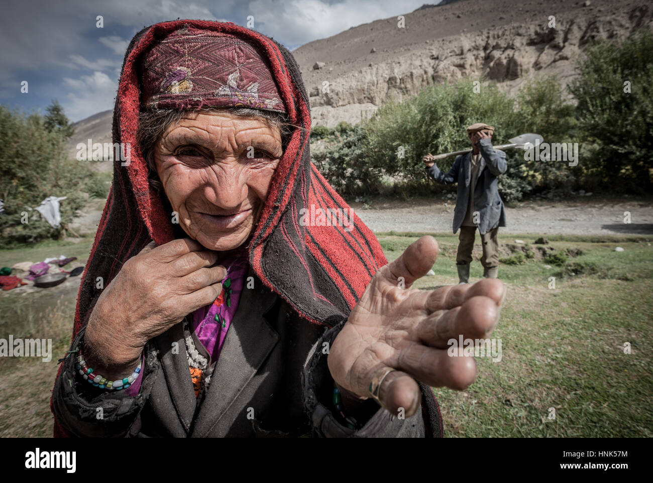 Afghanistan, Wakhan-Korridor, ein Porträt einer Frau mit roten Schleier im Tal, im Hintergrund ein Mann mit Schaufel. Stockfoto