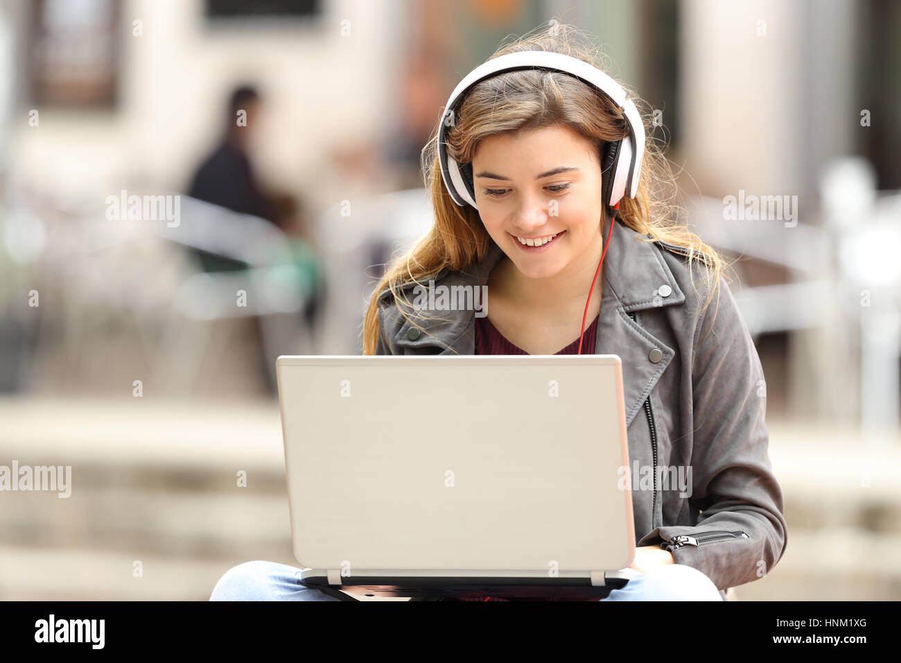 Casual Mädchen hören Musik mit Kopfhörern und Suche Lieder in einem Laptop sitzen auf einer Bank in der Straße Stockfoto