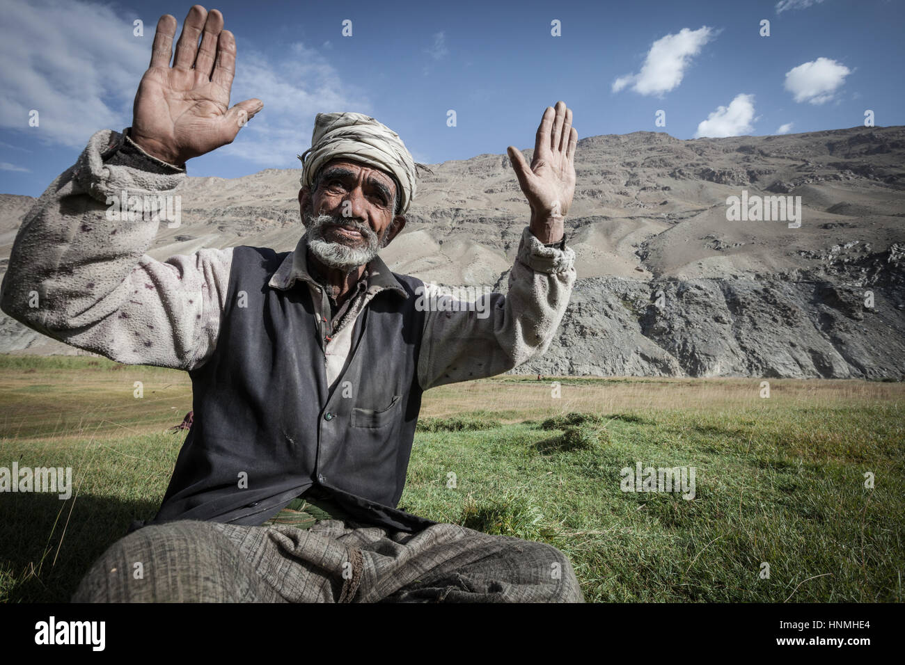 Afghanistan, Wakhan-Korridor, Alter Mann sitzt auf dem grünen Rasen auf dem Feld, sagt Hallo, Berge im Hintergrund. Stockfoto