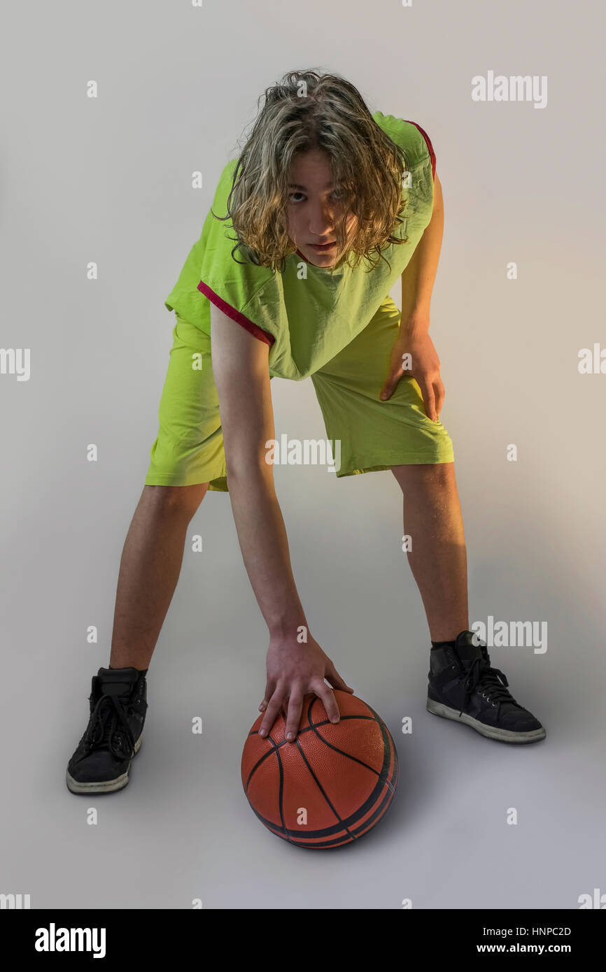 Kleiner Junge mit langen blonden Haaren, die mit einem grünen Trikot einen Basketball herunterdrücken und in die Kamera schaut Stockfoto