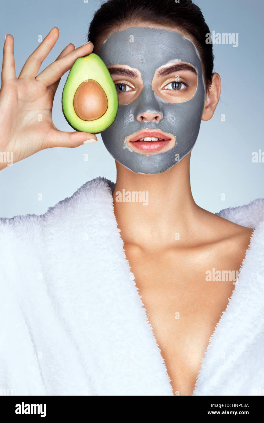 Hübsches Mädchen hält eine halbe Avocado vor ihr Gesicht. Foto eines Mädchens im weißen Bademantel und mit Maske aus Ton auf Gesicht. Pflege selbst Stockfoto