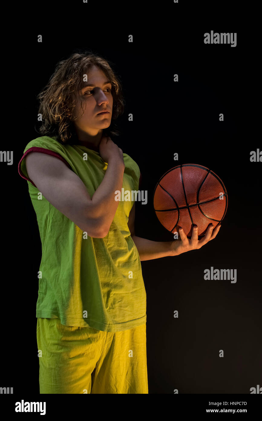 Junge mit langen blonden Haaren, die mit einem grünen Trikot streichelte seine Schulter und halten einen Basketball. Stockfoto