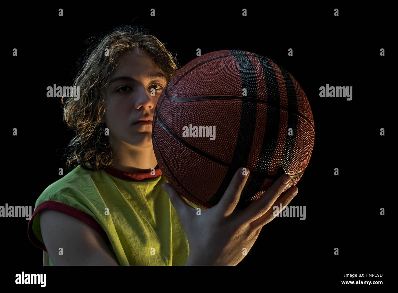 Nahaufnahme eines kleinen Jungen mit langen blonden Haaren, die mit einem grünen Trikot hält einen Basketball in die Kamera schaut. Stockfoto