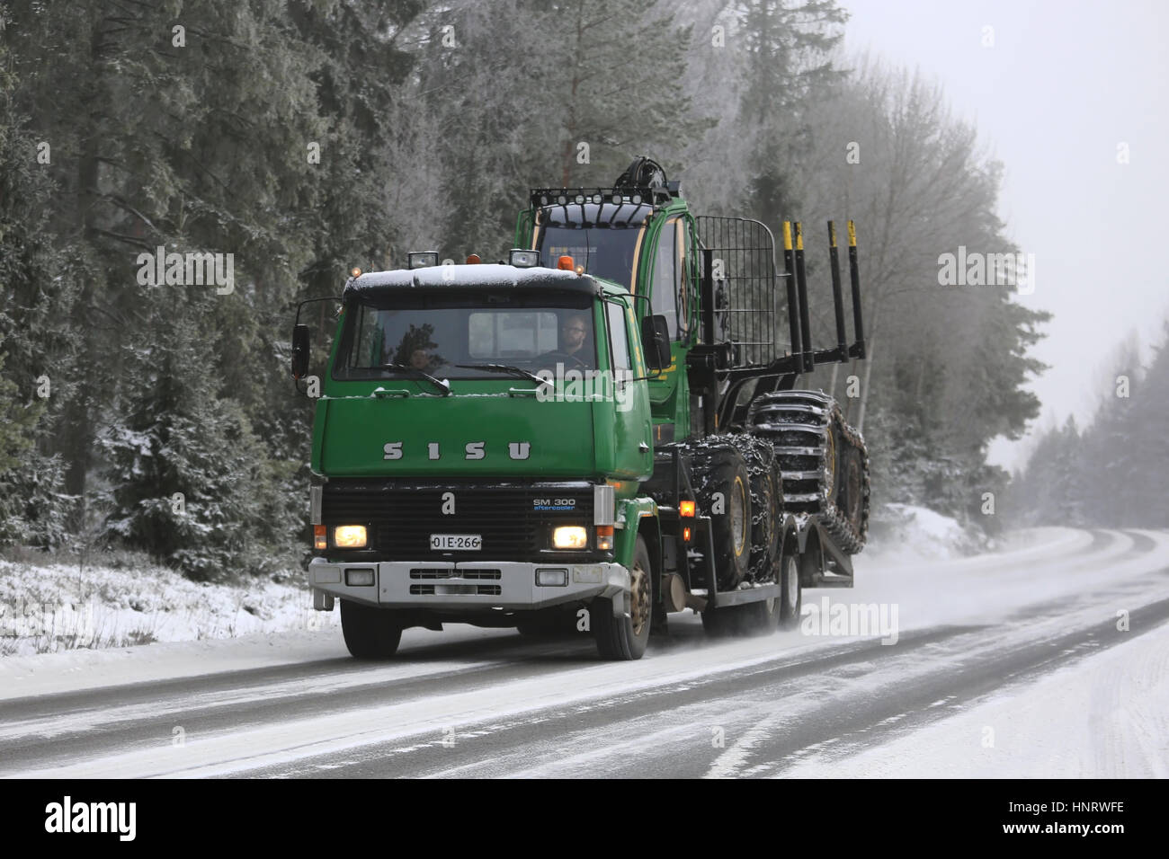 SALO, Finnland - 8. Januar 2017: Green SISU SM300 LKW transportiert ein John Deere Forstwirtschaft Spediteur an verschneiten Autobahn flankiert von Wald auf einer kalten win Stockfoto