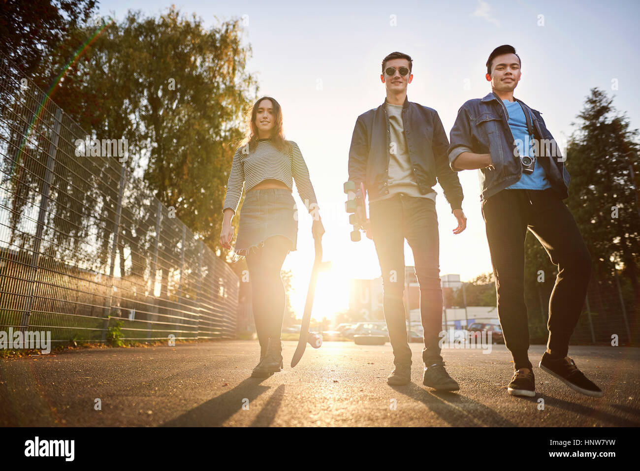 Porträt der jungen weiblichen Skateboarder und männlichen Freunde gehen auf der sonnendurchfluteten Straße Stockfoto