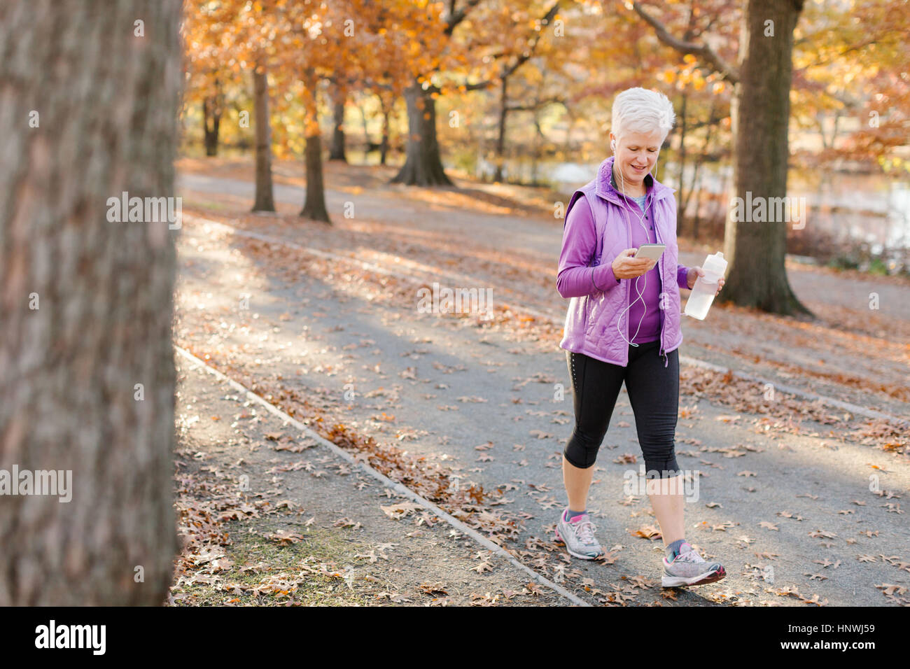 Senior Woman walking im Freien, mit Smartphone, trägt Kopfhörer und hält Wasser Flasche Stockfoto