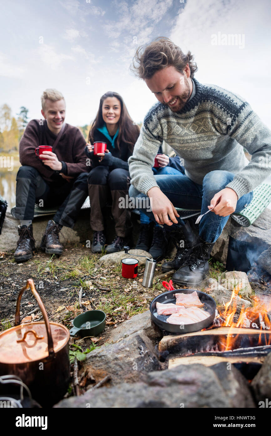 Mann Kochen am Lagerfeuer mit Freunden im Hintergrund Stockfoto