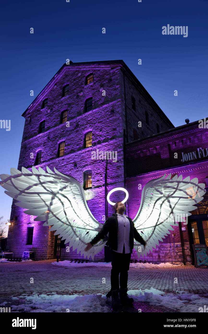 Mann mit leichten Halo und Engel Flügel an historischen Stein Mühlengebäude Distillery District Toronto Light Festival im Winter in der Abenddämmerung Stockfoto