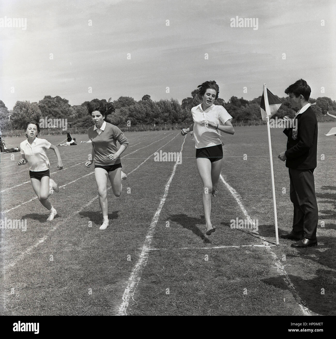 1960, historische, drei Mädchen, die an einem 100 Yard dash laufende Rennen auf Gras, mit Jungen in der Schule blazer Holding die Zeichenkette für die Ziellinie. Stockfoto