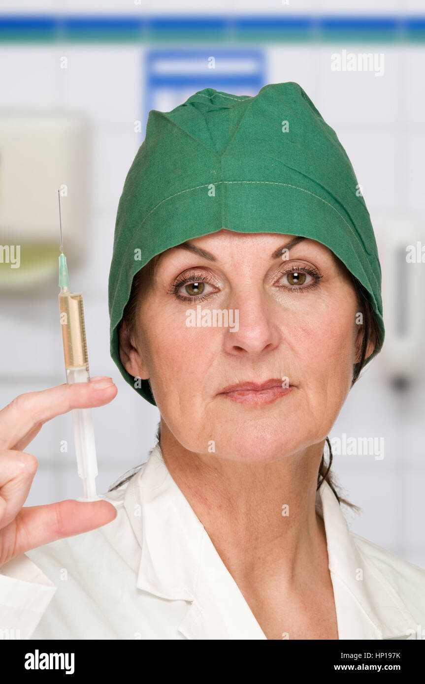 Kopf-Und-Schulter-Ansicht Einer Frau Mittleren Altersmit Grüner OP-Haube Vor Weißem Hintergrund Hält Eine Gefüllte Spritze Vor Sich. Stockfoto