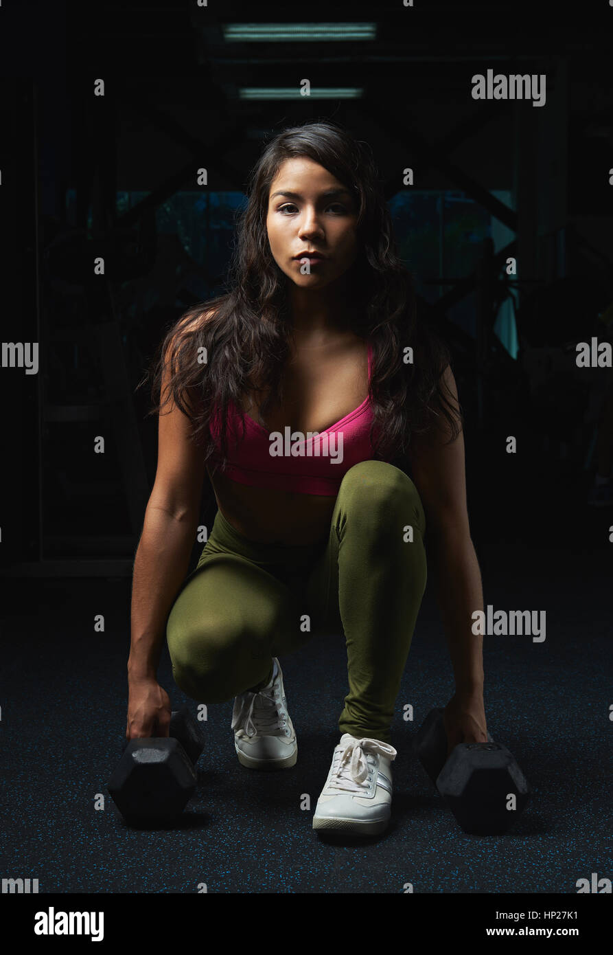 junge lateinamerikanische Frau sitzt mit Hanteln auf Fitness dunkle Turnhalle Hintergrund Stockfoto