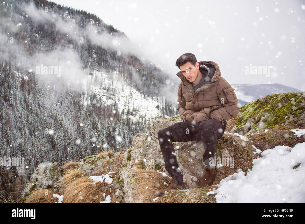 Hübscher junger Mann im Winter Oberbekleidung sitzen unter dem Schnee den Berg hinauf, beim Blick in die Kamera Stockfoto