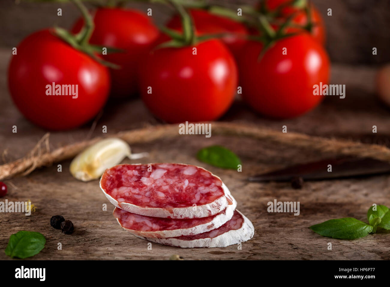 Italienische Salami-Scheiben auf Holz mit Cherry-Tomaten, Knoblauch und Kräutern im Hintergrund Stockfoto