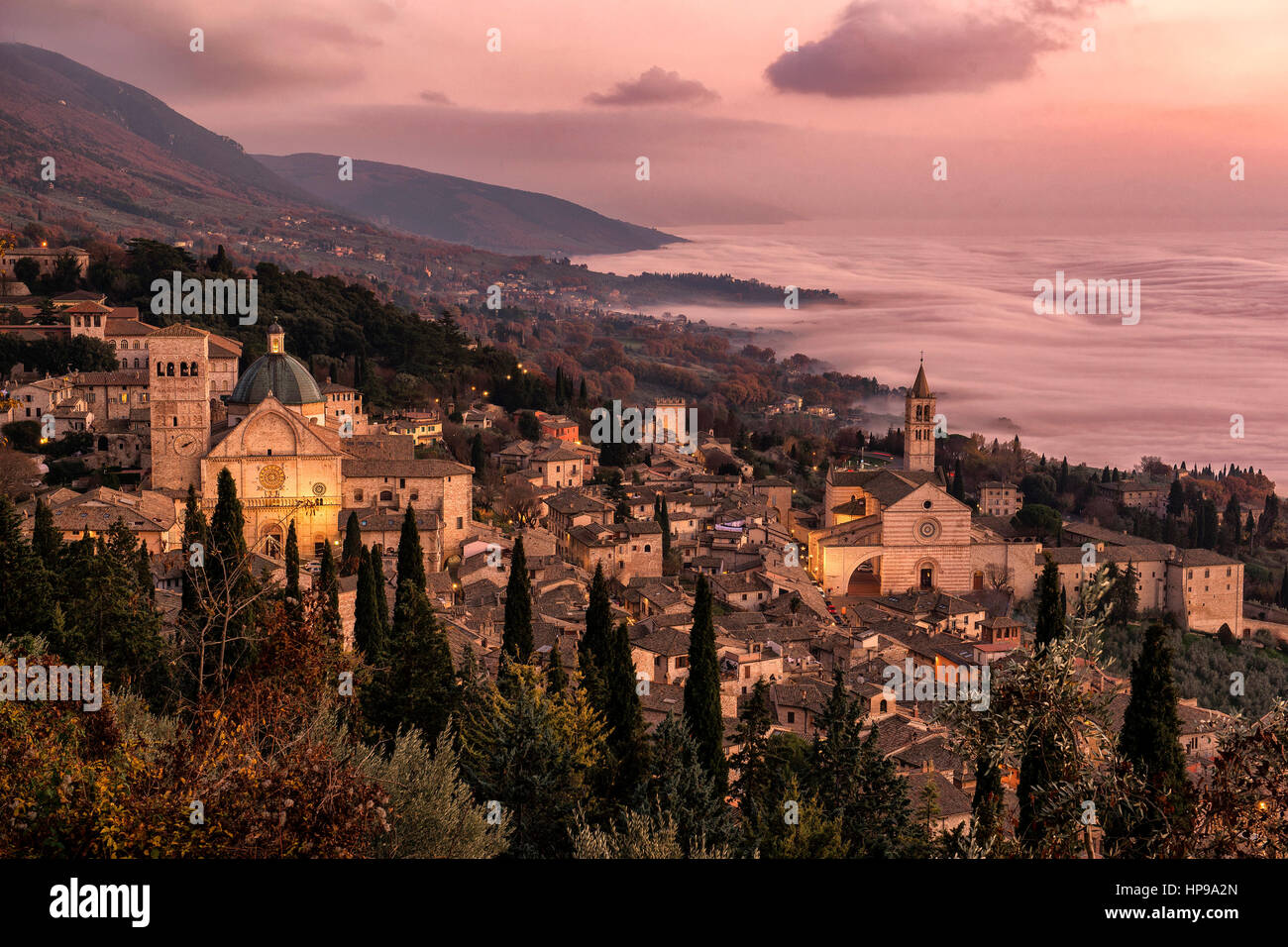 Assisi, Umbrien, Italien. Das Panorama von Assisi mit ihren Skyline mit der Glockentürme und Kirche von Saint Rufino und Saint Claire mit Nebel bei Sonnenuntergang Stockfoto